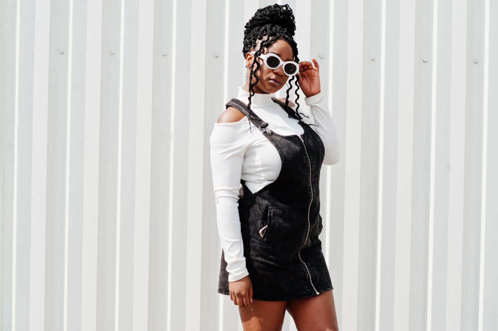 african woman in denim overalls skirt white sunglasses posed against white steel wall