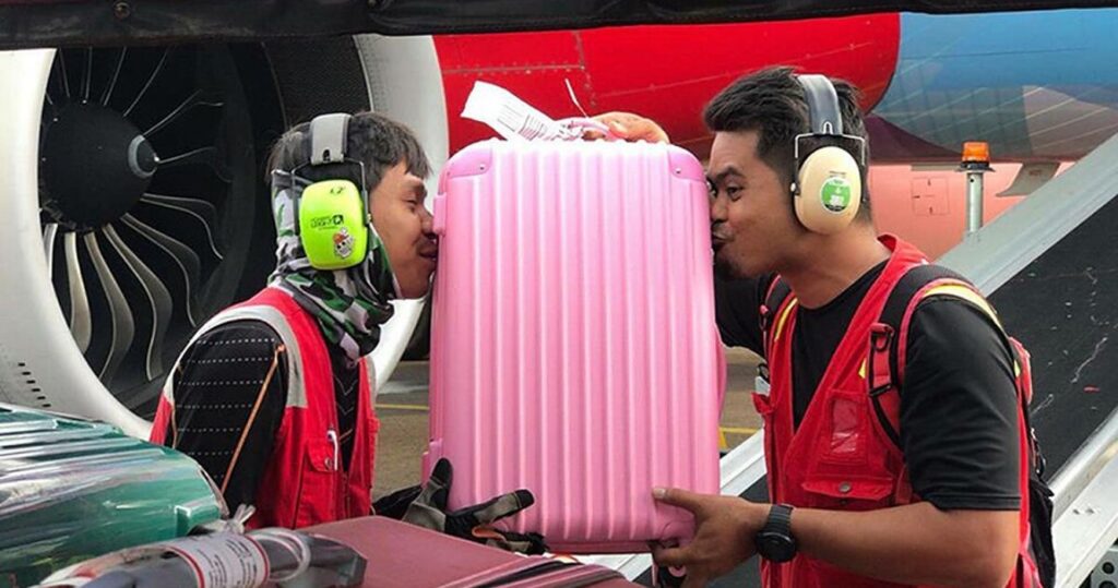 Two luggage handlers at the airport pretending to kiss someone's pink suitcase