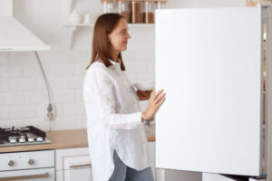 profile portrait beautiful young adult woman wearing white shirt looking smiling inside fridge with pleasant smile holding plate hands posing with kitchen set background